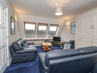 A living room with blue leather sofas a wooden coffee table a television and three large windows at Rothay 17 Ambleside