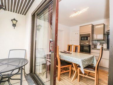 A dining area with wooden chairs and table covered with a tablecloth next to a kitchen with built-in oven and microwave visible through a glass door with outdoor furniture on the patio at Rothay 17 in Ambleside