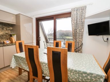 A dining area with a wooden table covered with a patterned cloth and six chairs next to sliding glass doors and kitchen cabinets at Rothay 17 in Ambleside