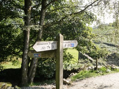 A wooden signpost on a gravel path with directions to Grasmere and Ambleside near trees and stone walls at Rothay 17 Ambleside