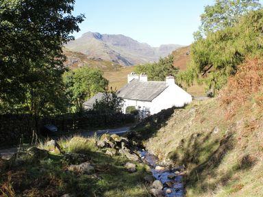A white house near a small stream with trees and hills in the background at Rothay 17 Ambleside