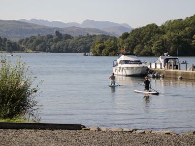 A lake with two people paddleboarding near boats and a dock with trees and mountains in the background at Rothay 17 in Ambleside