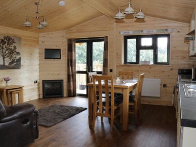 A wooden dining table with chairs in a wooden cabin room with a kitchen area a sofa and a wall-mounted television at Hayfields in Grimston