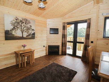 A wooden cabin room with nested tables a chair a wall-mounted TV a fireplace and glass doors leading outside at Hayfields in Grimston