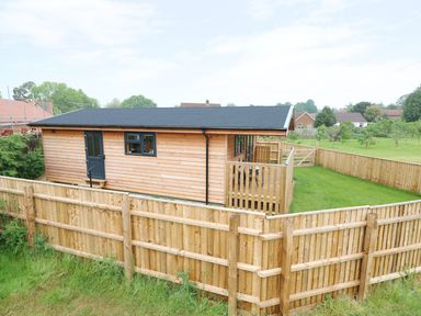 A wooden cabin with a black door and window surrounded by a wooden fence with green grass at Hayfields in Grimston