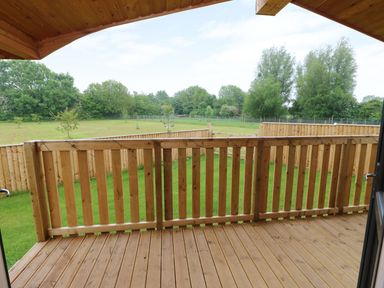 A wooden deck with railing overlooking a green lawn and fenced yard with trees at Hayfields Grimston