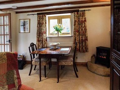 A dining area with a wooden table and four chairs next to a window with floral curtains and a small fireplace at The Old Cider House in Prestbury near Cheltenham