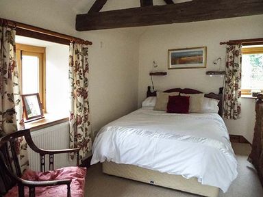 A bedroom with a double bed, floral curtains, wooden chair with cushion, two windows, and a framed picture on the wall at The Old Cider House in Prestbury near Cheltenham