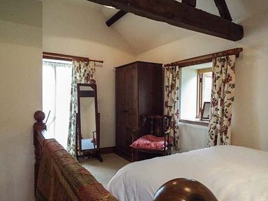 A bedroom with a wooden bed frame a standing mirror a wooden wardrobe a wooden chair with a cushion and floral curtains at The Old Cider House in Prestbury near Cheltenham