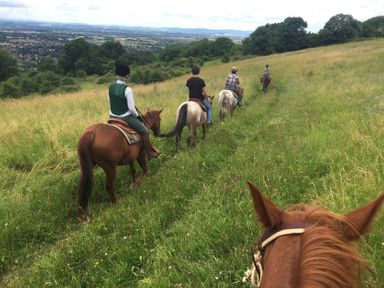 People riding horses on a grassy hillside with a view of a town and trees at The Old Cider House in Prestbury near Cheltenham