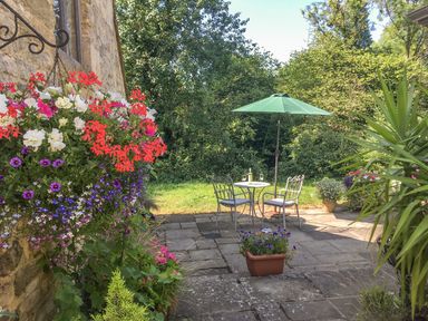 A garden patio with a small round table and two chairs under a green umbrella surrounded by green plants and colorful flowers at The Old Cider House in Prestbury near Cheltenham
