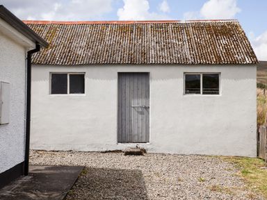 An exterior view of a white building with two windows and a central wooden door with a gravel ground at Shepherds Retreat in Gortin
