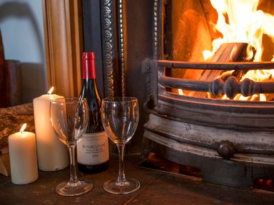 A bottle of red wine with two empty wine glasses and lit candles near a burning fireplace at Shepherds Retreat in Gortin