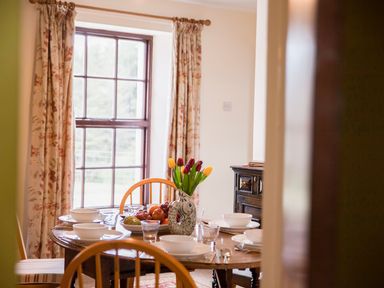 A dining room with a wooden table set with white bowls and plates a vase with tulips and a fruit bowl in front of a window with floral curtains at Shepherds Retreat in Gortin
