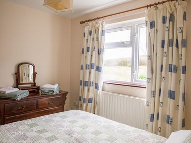 A bedroom with a bed covered in a patchwork quilt next to a wooden dresser with a mirror and folded towels by a window with patterned curtains at Shepherds' Retreat in Gortin