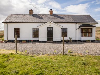 A white single-story house with a black door and windows surrounded by gravel and grass at Shepherds Retreat in Gortin