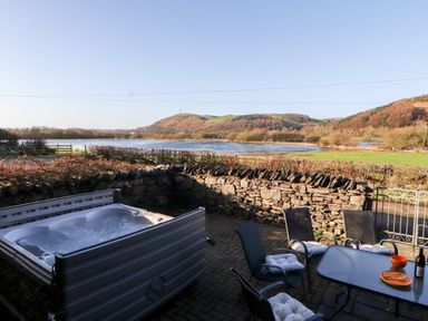 An outdoor patio with a hot tub and table with chairs overlooking a river and hills at Green Hills Farm in Ulverston