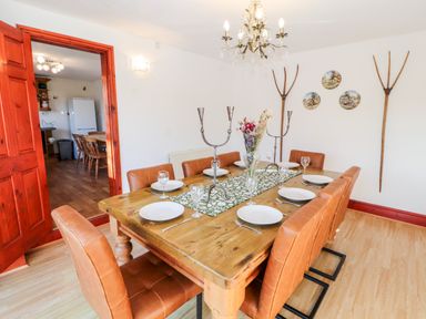 A dining room with a wooden table set with plates and glasses surrounded by brown chairs with a doorway leading to a kitchen at Green Hills Farm in Ulverston
