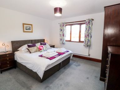 A bedroom with a double bed with pillows and folded towels bedside tables with lamps a window with floral curtains and a wooden wardrobe at Green Hills Farm in Ulverston