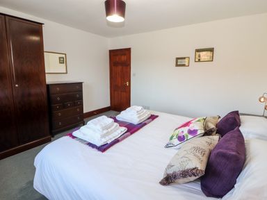 A bedroom with a white bed with purple and floral pillows a wooden wardrobe and chest of drawers and a wooden door at Green Hills Farm in Ulverston