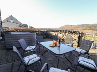 An outdoor patio with a glass table surrounded by six cushioned chairs and a stone wall in the background at Green Hills Farm in Ulverston