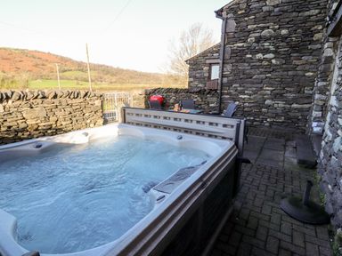 An outdoor hot tub on a stone patio next to a stone building with outdoor chairs and a table at Green Hills Farm in Ulverston