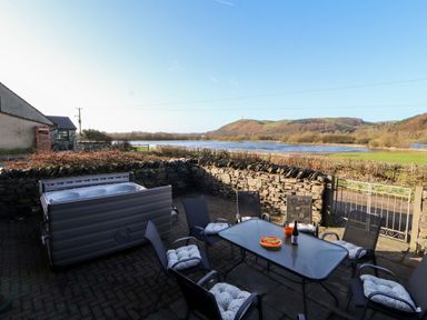 An outdoor patio with six chairs around a table and a hot tub next to a stone wall overlooking a river and hills at Green Hills Farm in Ulverston