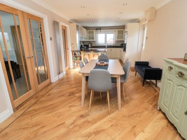 A dining room with a table and six chairs near a kitchen with white cabinets and wooden flooring at The Croft in Longhoughton near Alnwick