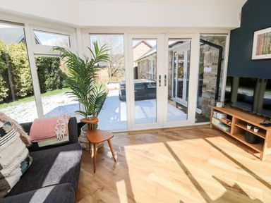 A living room with a dark sofa with cushions and blankets a wooden side table with a plant a tv on a wooden stand and glass doors showing a snowy garden at The Croft in Longhoughton near Alnwick