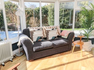 A sunlit seating area with a gray upholstered sofa decorated with patterned cushions and throws near windows and a wooden side table with a plant at The Croft in Longhoughton near Alnwick