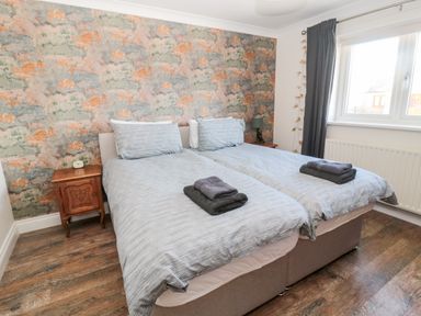 A bedroom with two single beds side by side with gray bedding and folded towels on top a wooden nightstand with an alarm clock against a wall with patterned wallpaper and a window with gray curtains at The Croft in Longhoughton near Alnwick