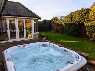 An outdoor hot tub on a wooden deck beside a house with glass doors and a garden with grass and bushes