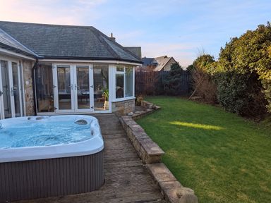 A backyard with a hot tub wooden deck grass lawn bushes and a house with glass doors at The Croft in Longhoughton near Alnwick