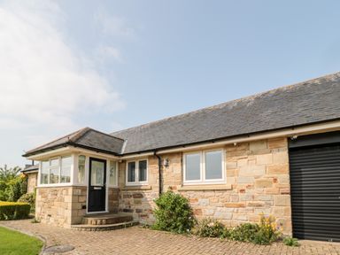 The front exterior of a single-story stone house with a black door, windows, a garage door, and a paved driveway with plants along the side at The Croft in Longhoughton near Alnwick