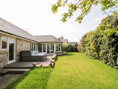 A backyard with a wooden deck and a hot tub next to a stone house with glass doors and windows at The Croft in Longhoughton near Alnwick