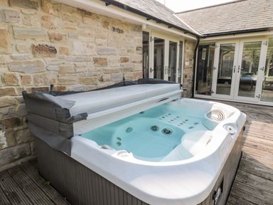 An outdoor hot tub with a partially open cover on a wooden deck next to a stone wall and glass doors at The Croft in Longhoughton near Alnwick