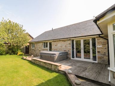 An outdoor area with a wooden deck and hot tub next to a stone house and grass lawn at The Croft in Longhoughton near Alnwick