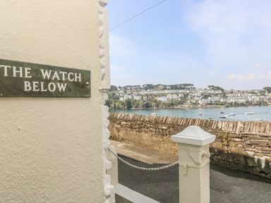 A street view with a wall and a sign that reads The Watch Below overlooking a body of water and houses on a hillside at The Watch Below in Polruan