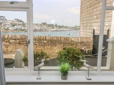 A window with two bird figurines and a small plant on the sill overlooking a stone wall a water body with boats and houses in the background at The Watch Below in Polruan