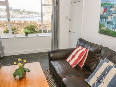 A living room with a dark leather sofa with striped cushions and a wooden table with flowers in a pot by a window showing a seaside town at The Watch Below in Polruan