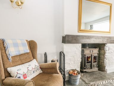 An armchair with a blue and white blanket and a cushion next to a fireplace with firewood and a wood burner at The Watch Below in Polruan