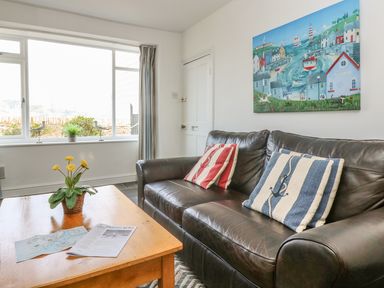 A living room with a leather sofa two striped cushions a wooden coffee table with flowers and papers and a colorful painting on the wall at The Watch Below in Polruan
