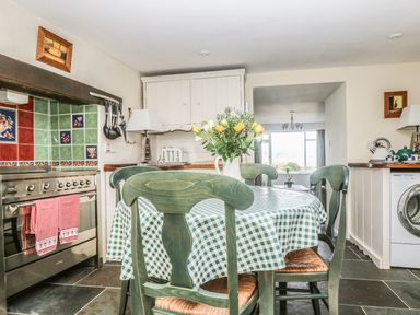 A kitchen with a dining table covered with a checkered tablecloth and chairs a stove with red and green tiled backsplash and a washing machine at The Watch Below in Polruan