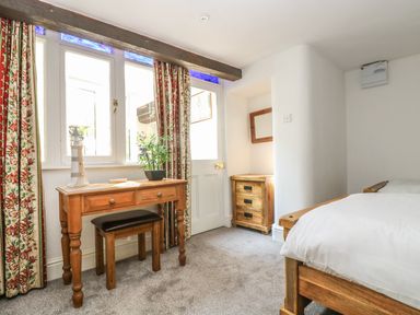 A bedroom with a wooden desk and stool near window and door with floral curtains a wooden bedside table a bed with white bedding at The Watch Below in Polruan
