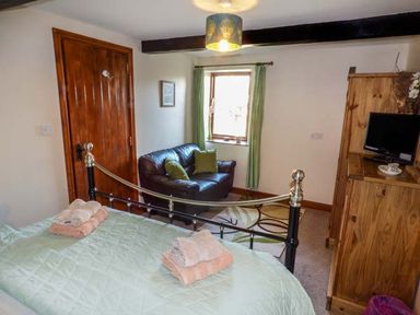 A bedroom with a bed, towels on the bed, a brown leather sofa, a wooden wardrobe with a TV, and a window with green curtains at Bank End Barn in Grizebeck