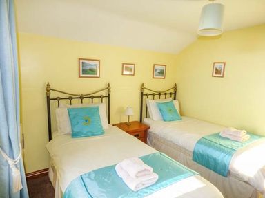 A bedroom with two single beds with blue cushions and throws a wooden nightstand a lamp and framed pictures on a yellow wall at Bank End Barn in Grizebeck