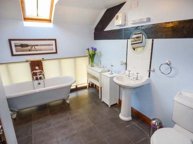 A bathroom with a clawfoot bathtub under a skylight a pedestal sink a toilet a small cabinet and a vanity table with flowers at Bank End Barn in Grizebeck