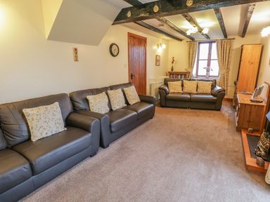 A living room with two brown leather sofas with floral cushions a wooden cabinet a window with curtains and a wall clock at Bank End Barn in Grizebeck