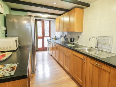 A narrow kitchen with wooden cabinets a black countertop a stainless steel sink a microwave a fridge and wooden floor at Bank End Barn in Grizebeck
