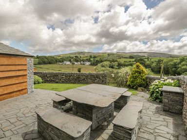 An outdoor patio area with stone table and benches overlooking a stone wall and green fields at Bank End Barn in Grizebeck